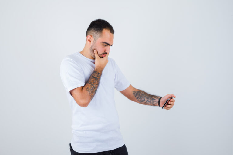 young man looking at something on phone and putting hand on chin in white t-shirt and black pants and looking serious. front view